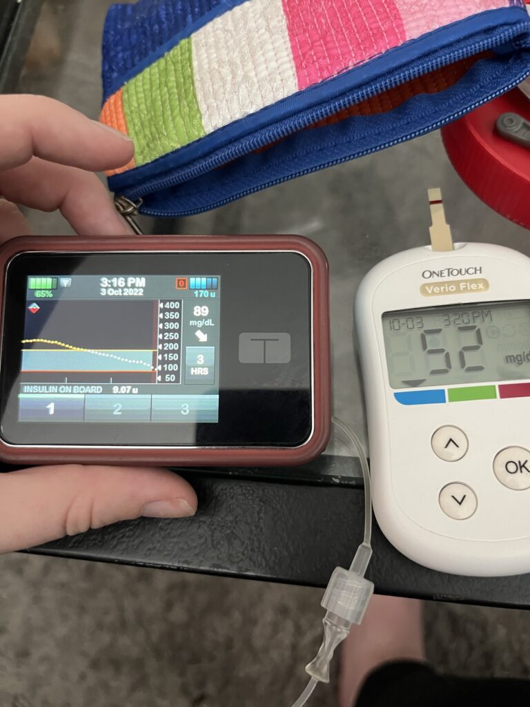 Clara's hand holding her insulin pump. which shows a blood sugar reading of 89. Her glucometer is on a glass table with a blood sugar reading of 52.